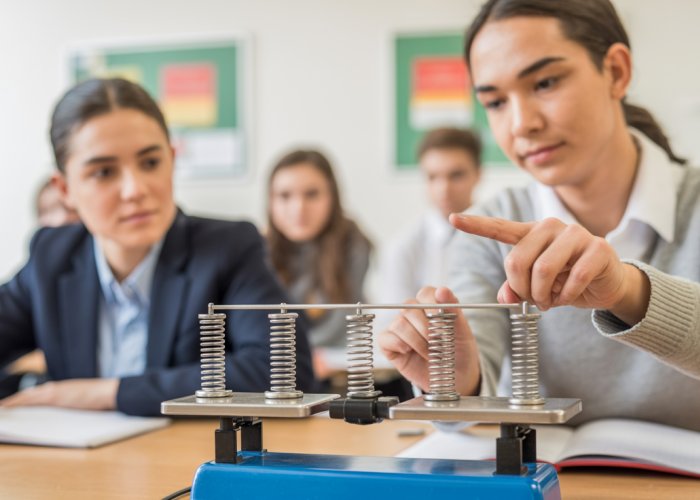 Teacher demonstrating the relationship between kilograms and Newtons using a spring scale and weights in a physics classroom.